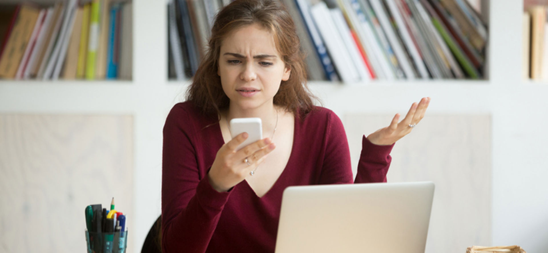 woman looking at phone frustrated while sitting behind a laptop at a desk